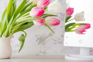 A bouquet of white and pink tulips with green leaves stands in a jug on the table and is reflected in the mirror. Selective focus. photo