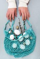 Happy Easter. Female hands hold a blue string bag with chicken and quail eggs and pussy willow branches on a gray background photo