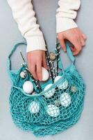 Happy Easter. Female hands put chicken and quail eggs and pussy willow branches in a blue string bag on a gray background photo