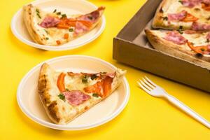 Takeaway and delivery. Pizza slice in disposable plastic plate, pizza box and fork on yellow background photo