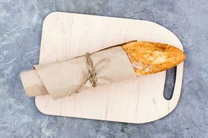 Fresh baguette in wrapping paper on cutting board on table. Top view photo