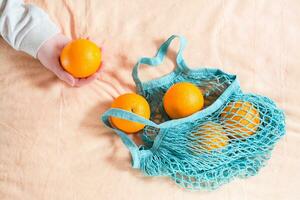 A female hand pulls out fresh oranges from a mesh bag on a fabric background. Zero waste photo