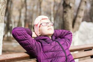 A girl in a jacket and glasses on a bench in a city park in early spring put her face to the rays of the sun and enjoys the first warmth photo