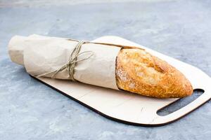 Fresh baguette in wrapping paper on cutting board on table photo
