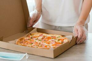 para llevar alimento. un mujer en desechable guantes sostiene un cartulina caja de Pizza y un protector máscara en el mesa en el cocina foto