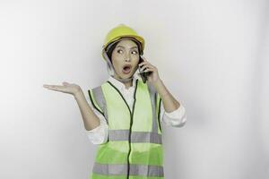 A smiling Asian woman labor wearing safety helmet and vest, pointing to copy space beside her while having a phone call, isolated by white background. Labor's day concept. photo