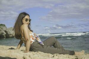 joven asiático mujer sentar en el playa arena. retrato sexy asiático dama de viaje y relajante en el verano con tropical naturaleza. foto