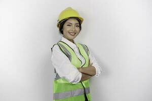 Smiling Asian woman labor worker in industry factory, posing with arms folded, wearing yellow safety helmet, green vest and uniform, isolated white background. photo