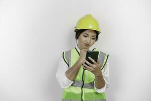 A thoughtful young woman labor worker wearing safety helmet and vest while holding her phone and hand on her chin, isolated by white background. Labor's day concept. photo