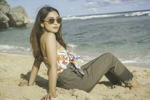 joven asiático mujer sentar en el playa arena. retrato sexy asiático dama de viaje y relajante en el verano con tropical naturaleza. foto