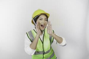 Young beautiful woman labor wearing safety helmet and vest is shouting and screaming loud with a hand on her mouth. Labor's day concept. photo