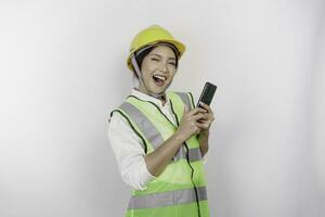 un sonriente asiático mujer labor vistiendo la seguridad casco y chaleco mientras participación su teléfono inteligente, aislado por blanco antecedentes. mano de obra día concepto. foto
