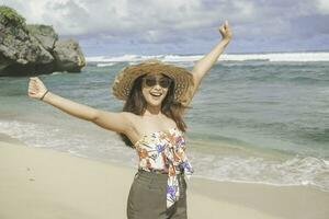 A happy young Asian woman wearing beach hat is posing to the camera with her hands open, expressing freedom and carefree holiday at the beach in Gunungkidul, Indonesia photo