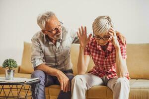 Senior couple sitting in living room and arguing. Woman is angry while man is apologizing. photo