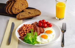 Hearty breakfast with scrambled eggs, bacon, beans, cherry tomatoes, homemade bread, orange juice on a concrete countertop. English breakfast concept. photo