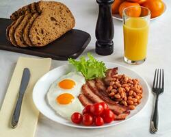 Hearty breakfast with scrambled eggs, bacon, beans, cherry tomatoes, homemade bread, orange juice on a concrete countertop. English breakfast concept. photo