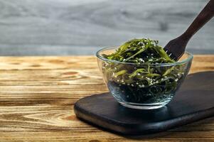 Seaweed salad. Asian cuisine. Seaweed kelp in a glass bowl sprinkled with sesame seeds, on a wooden board with a wooden fork. Laminaria photo