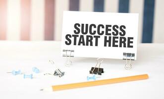 SUCCESS START HERE sign on paper on white desk with office tools. Blue and white background photo