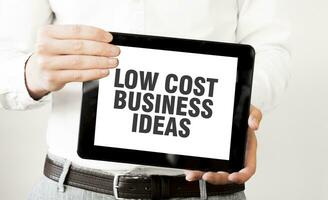 Text LOW COST BUSINESS IDEAS on tablet display in businessman hands on the white background. Business concept photo