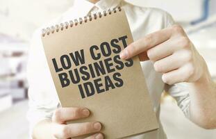 Text LOW COST BUSINESS IDEAS on brown paper notepad in businessman hands in office. Business concept photo