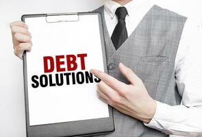 DEBT SOLUTIONS inscription on a notebook in the hands of a businessman on a gray background, a man points with a finger to the text photo