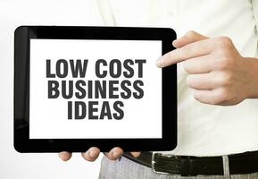 Text LOW COST BUSINESS IDEAS on tablet display in businessman hands on the white background. Business concept photo