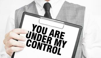 YOU ARE UNDER MY CONTROL inscription on a notebook in the hands of a businessman on a gray background, a man points with a finger to the text photo