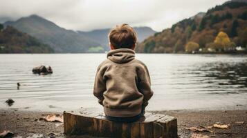 a boy sitting on a log by a lake with mountains in the background generative ai photo