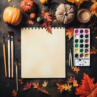 Notebook template on halloween background. White square sketchbook lies on brown wooden table with paints, brushes, halloween pumpkins, orange leaves. Halloween background. Minimalist product mockup. photo