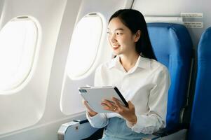 Attractive Asian female passenger of airplane sitting in comfortable seat while working laptop and tablet with mock up area using wireless connection. Travel in style, work with grace. photo