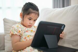 Asian child girl using pen and touch drawing on tablet display screen. Baby smiling funny time to use tablet. Too much screen time. Cute girl watching videos while tv, Internet addiction concept. photo