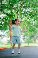 Asian child girl is jumping on trampoline on playground background. Happy laughing kid outdoors in the yard on summer vacation. Jump high on trampoline. Activity children in the kindergarten school. photo