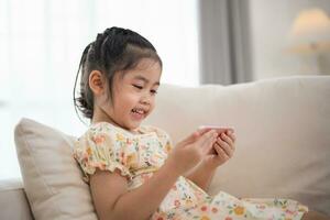 Asian child girl looking using and touch mobile phone screen on couch sofa. Baby smiling funny time to use mobile phone. Too much screen time. Cute girl watching videos while tv, Internet addiction. photo