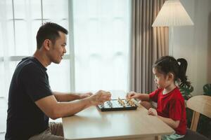 Education studying concept. Little cute asian baby girl children playing chess with her father dad in the living room at home.Smart kid. Little genius Child. Intelligent game.Chessboard. photo