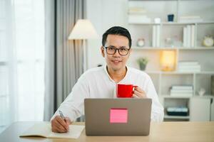 inteligente asiático hombre sonriente vistiendo lentes trabajando con computadora ordenador portátil. concepto trabajo formar hogar, permanecer a hogar. Lanza libre vida estilo, nuevo normal social distanciamiento estilo de vida. trabajo formar en cualquier sitio concepto. foto