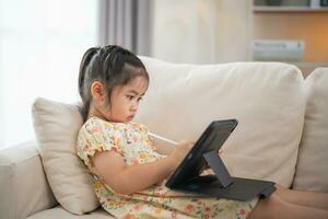 Asian child girl using pen and touch drawing on tablet display screen. Baby smiling funny time to use tablet. Too much screen time. Cute girl watching videos while tv, Internet addiction concept. photo