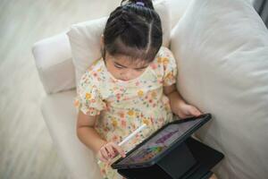 Asian child girl using pen and touch drawing on tablet display screen. Baby smiling funny time to use tablet. Too much screen time. Cute girl watching videos while tv, Internet addiction concept. photo