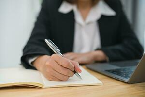 Close up of handAsian businesswoman wearing suit and writing notes book and working laptop on table at home. Entrepreneur woman working for business at living room home. Business work home concept. photo