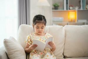 alegre contento asiático niño bebé niña sonriente y leyendo libro mientras sentado en sofá sofá en vivo habitación a hogar. niña relex leyendo libro sonrisa a sofá en el casa. espalda a colegio concepto. foto