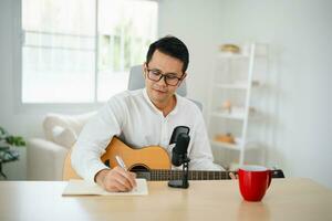 asiático hombre vistiendo auricular jugar guitarra y canta un canción y aprendizaje en línea con ordenador portátil y cuaderno y componer escritura canción, grabar sonido y utilizar computadora grabación música programa. músico concepto. foto