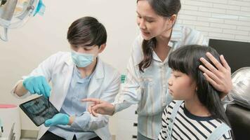 asiático masculino pediátrico dentista explica dientes radiografía escanear a niña y su madre para odontología higiene desde tableta en dental clínica, profesional ortodoncia oral médico en un niño cuidado de la salud hospital. video