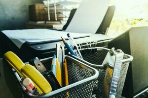 Close up of stationary items such as pencils, scissors, cutter knives, and paper clips are placed in a stainless steel basket and placed on table by office window. Concept of equipment used in work. photo