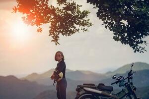 woman holding safety helmet standing beside enduro motorcycle against beautiful mountain scene of khaoyai national park thailand photo