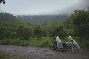 asiático parejas colaboró a empujar un mal funcionamiento motocicleta en el medio de el bosque la carretera foto