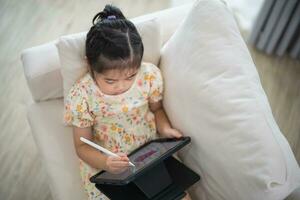 Asian child girl using pen and touch drawing on tablet display screen. Baby smiling funny time to use tablet. Too much screen time. Cute girl watching videos while tv, Internet addiction concept. photo