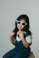 Portrait of relaxed cute little asian girl child funny smiling and laughing wearing white headphones and glasses, keeps hands on her ears, posing and smiling enjoying isolated over gray background. photo