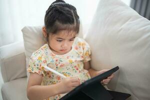 Asian child girl using pen and touch drawing on tablet display screen. Baby smiling funny time to use tablet. Too much screen time. Cute girl watching videos while tv, Internet addiction concept. photo