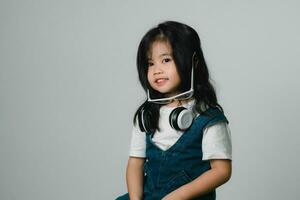 retrato de relajado linda pequeño asiático niña niño gracioso sonriente y riendo vistiendo blanco auriculares y anteojos, mantiene manos en su orejas, posando y sonriente disfrutando aislado terminado gris antecedentes. foto