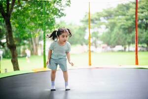 asiático niño niña es saltando en trampolín en patio de recreo antecedentes. contento riendo niño al aire libre en el yarda en verano vacaciones. saltar alto en trampolín. actividad niños en el jardín de infancia escuela. foto