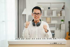 Asian man wearing white headphones play piano and sing a song and learning online with mobile phone and compose writing song or record sound with a microphone and use computer recording music program photo